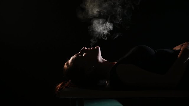 The brunette smokes an electronic cigarette on a black background, releases thick smoke from her mouth. Portrait of a woman smoking a vape, hovering.