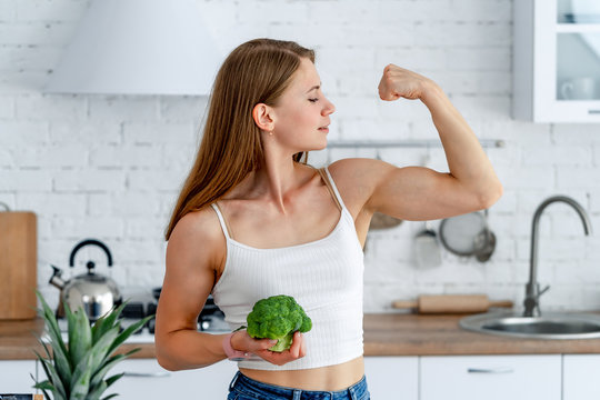 Strong woman with broccoli in the kitchen. Young healthy girl shows her strong hands.
