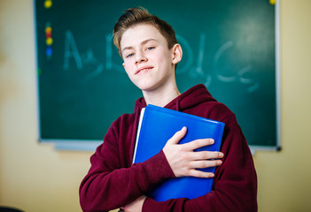 Student of higer school is standing in dark hoodie near the green blackboard in the classroom. Teenage Boy with his blue notebook in hands.