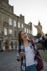 Young cheerful woman with happy emotional face waiting for taxi outdoors. Girl with backpack walking to university, using mobile phone, listening music, dancing, laughing on urban street 