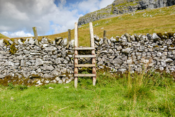 Detailed view of a wooden, public right of way structure to climb over a stone wall. An empty road leads to the summit of the mountain.
