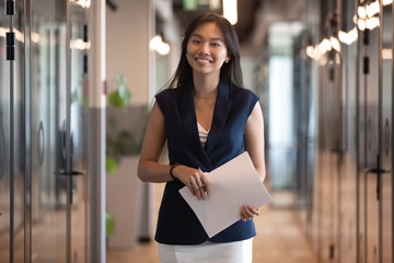 Portrait of smiling Asian businesswoman holding document, standing in hallway - obrazy, fototapety, plakaty