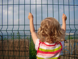 Blond child is holding on to a metal fence. The view from the back. Concrete blocks. Clouds...