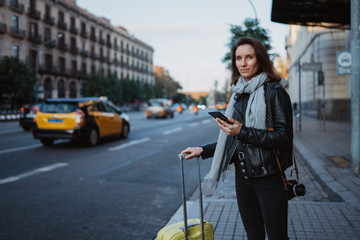 Traveler woman with suitcase calling mobile phone waiting yellow taxi in evening street europe city...
