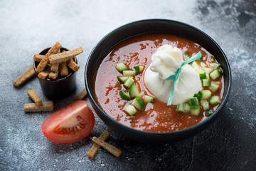 Black bowl of gazpacho soup with burrata cheese and chopped cucumber, studio shot on a grey stone background, selective focus