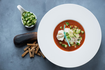 White plate with tomato gazpacho, burrata and chopped cucumber, flatlay over grey concrete background