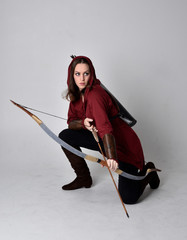 full length portrait of a brunette girl wearing a red fantasy tunic with hood, holding a bow and...