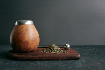 Calabas and a bombilla with a bunch of mate tea on a wooden plate on dark background. Organic traditional tea. Free space for your text.