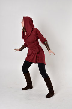 full length portrait of a brunette girl wearing a red fantasy tunic with hood. Standing pose in side profile on a white studio background.