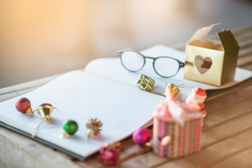 Book note with Christmas decoration on wooden table and flare light blurred background. Ball,Gift box ,Santa clause,Branch pine,bell, glasses.Blurred foreground.