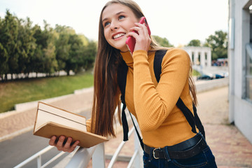 Attractive smiling student girl with notebook happily talking on cellphone studying in park