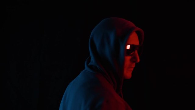 Adult man in black hood and sunglasses turning and looking to camera on black background. Unrecognizable man in black hoodie and glasses in blue and red lighting in dark studio