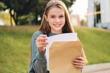 Beautiful cheerful casual student girl opening envelope with exams results happily looking in...