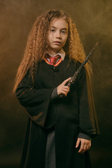 Portrait of a little witch girl with long brown hair, dressed in dark coat, holding magic wand in her hand, posing on black smoky studio background.