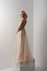 Girl in a dress on a white background standing on a white cube. Dress with rhinestones and beads. Hands on hips. Hairstyle and makeup.