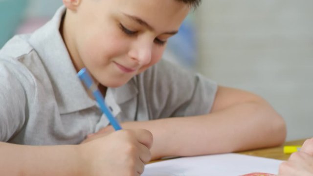 Tilt up of cute little boy smiling and talking with female teacher while drawing with colored pencil at table in kindergarten