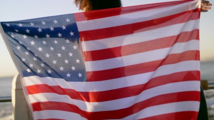 African american woman holding USA flag by the ocean, celebrating independence day sunflare at sunset