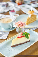Piece of delicious cheesecake with strawberry and mint leaves on white plate. Breakfast in the cafe, morning coffee. Cappuccino and lots of desserts on the table.