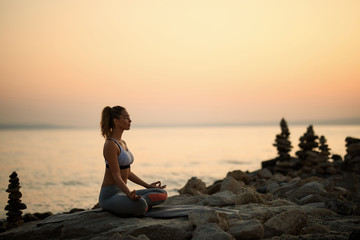 Zen-like woman meditating on a rock by the sea at dusk.