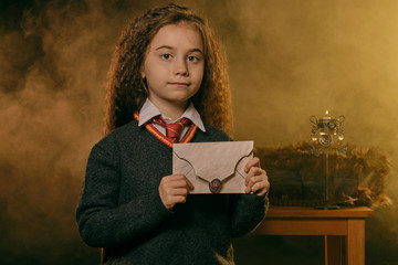 Little witch girl dressed in dark clothes, standing near a table and holding a letter in her hands while posing against a black smoky background.