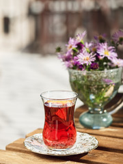 Tea in a glass of armudu on a saucer, located on a wooden table in the loggia. In the background are flowers . Bright sunny day. Close-up.