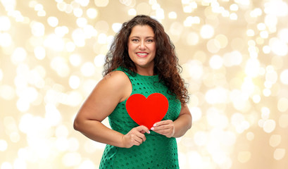 valentine's day, holidays and love concept - happy woman in green dress holding red heart over festive lights on beige background