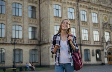 Young attractive student with backpack walking to university, education concept. Beautiful hipster woman holding mobile phone, listening to music, laughing  
