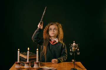 Little witch dressed in dark clothes sitting at the table against a black smoky background. There are magic wand, books with spells, candles on it.