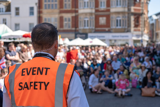 Shallow focus of an Event Safety officer seen facing members of the public in a town square. The public are watching the Royal Wedding on a large, outside screen.
