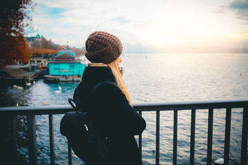 Young beautiful pretty blonde tourist girl in warm hat and coat with backpack walking at cold autumn in Europe city enjoying her travel in Zurich Switzerland on sunset at lake