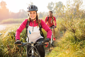 Beautiful happy young woman riding mountain bike over a meadow by the lake or river in the early morning. Young man blurred in the background. Healthy and active sporty lifestyle