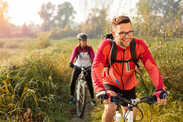 Beautiful happy young man riding mountain bike over a meadow by the lake or river in the early morning. Young woman blurred in the background. Healthy and active sporty lifestyle