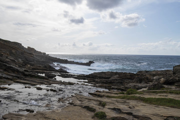 Fototapeta na wymiar Orilla rocosa con olas en el mar y cielo azulado nuboso.