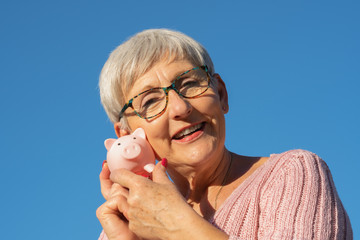 smiling older woman with White hair, glasses, pink sweater and White teeth with a piggy bank in hands on isolated blue background