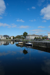 Galway city on a sunny summer day - taken at Eglington Basin near the Spanish Arch showing surrounding buildings & trees, with sailboats moored on calm reflective water.