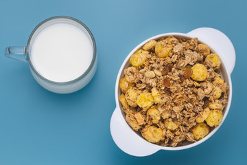 A mixture of cereals dried and nuts with milk at breakfast. Glass cup of milk with rolled oats in a white plate on blue table. White bowl of granola with glass of milk on blue background.