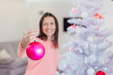 Young woman in a pink sweater holding a Christmas decoration bauble for the Christmas tree, showing...