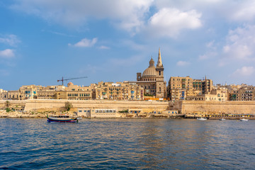 Fototapeta na wymiar Coastal landscape of Valletta at sunny summer day, with boat drifting in the foreground. Maltese Valletta skyline with church of Our Lady of Mount Carmel and St Paul's Pro-Cathedral.