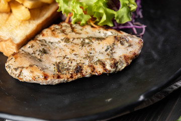 Grilled chicken steak served with french fries, garlic bread and mixed salads in a black matt plate on a real wood background. Chicken for all can eat, good taste, easy cook. Good food for good health