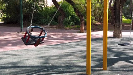 Slow motion shot of an empty swing, swinging alone in a playground without children. The trees in the background cast shadows over the new safety matt and the old brick road. Steady handled.
