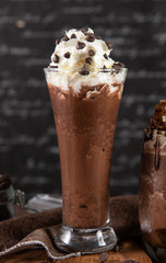 Ice chocolate, toppings by whipping cream and choc chip in a tall of clear glass on dark gray real wood and chopping board. Decorat with choc chip on floor. Popular drink to quench thirst to cool down