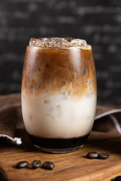 Ice coffee, toppings by floating foam milk and burned sugar in a tall of clear glass on dark gray real wood and chopping board. Decoration with coffee bean. Popular drink to quench thirst to cool down