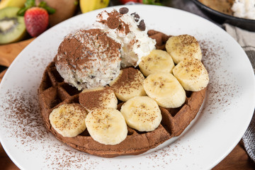 The banana chocolate waffle, topping with whipped cream and ice cream on white plate. Decorated with banana and chocolate chip on chopping real wood background. Eat sweet in moderation make to happy