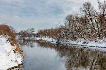 Small river in winter at sunny day against cloudy sky. Country winter landscape