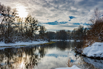 Fototapeta premium River landscape in sunny winter day. Snow covered riversides of small river with trees and bushes in sunlight against blue sky with clouds