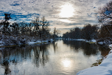 Winter river landscape. Sun and blue sky with clouds reflected on the water surface of river against snow covered riversides