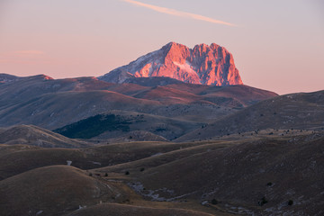 Mountain Corno Grande on horizon behind barren and rural landscape glowing pink in light of sunrise, Abruzzo, Italy