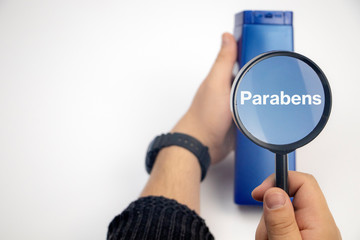 Chemical components on the shampoo label: parabens . A hand holds a blue jar and a magnifier, where...