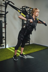 Beautiful blonde woman wearing in sensors and belts, fastens with velcro is practicing EMS fitness in a gym performing traction exercises.
