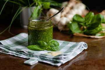 Organic homemade traditional italian sauce pesto for pasta in glass jar: leaves of basil, cheese, nuts and olive oil. Dark and moody, low key, closeup. Cozy atmosphere 